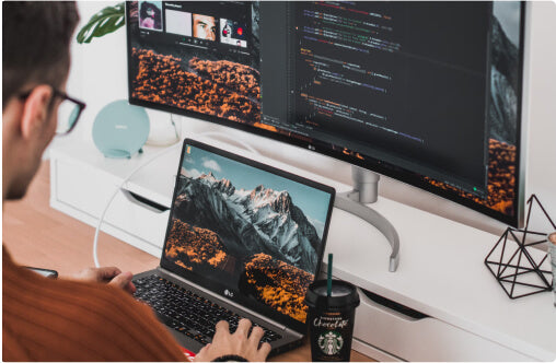 A white male at a desk, working on a laptop connected to a large display screen behind it