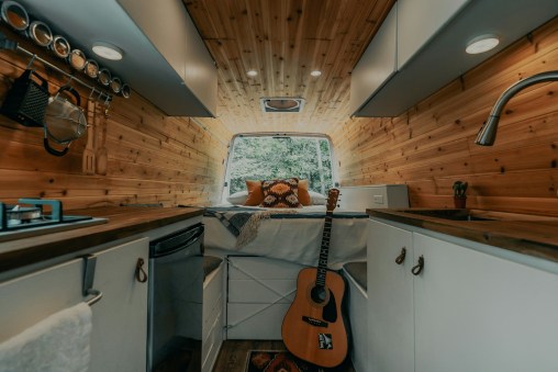 View into the back of a camping vehicle showing wooden clad ceiling, kitchen units, appliances, lights, guitar and a bed