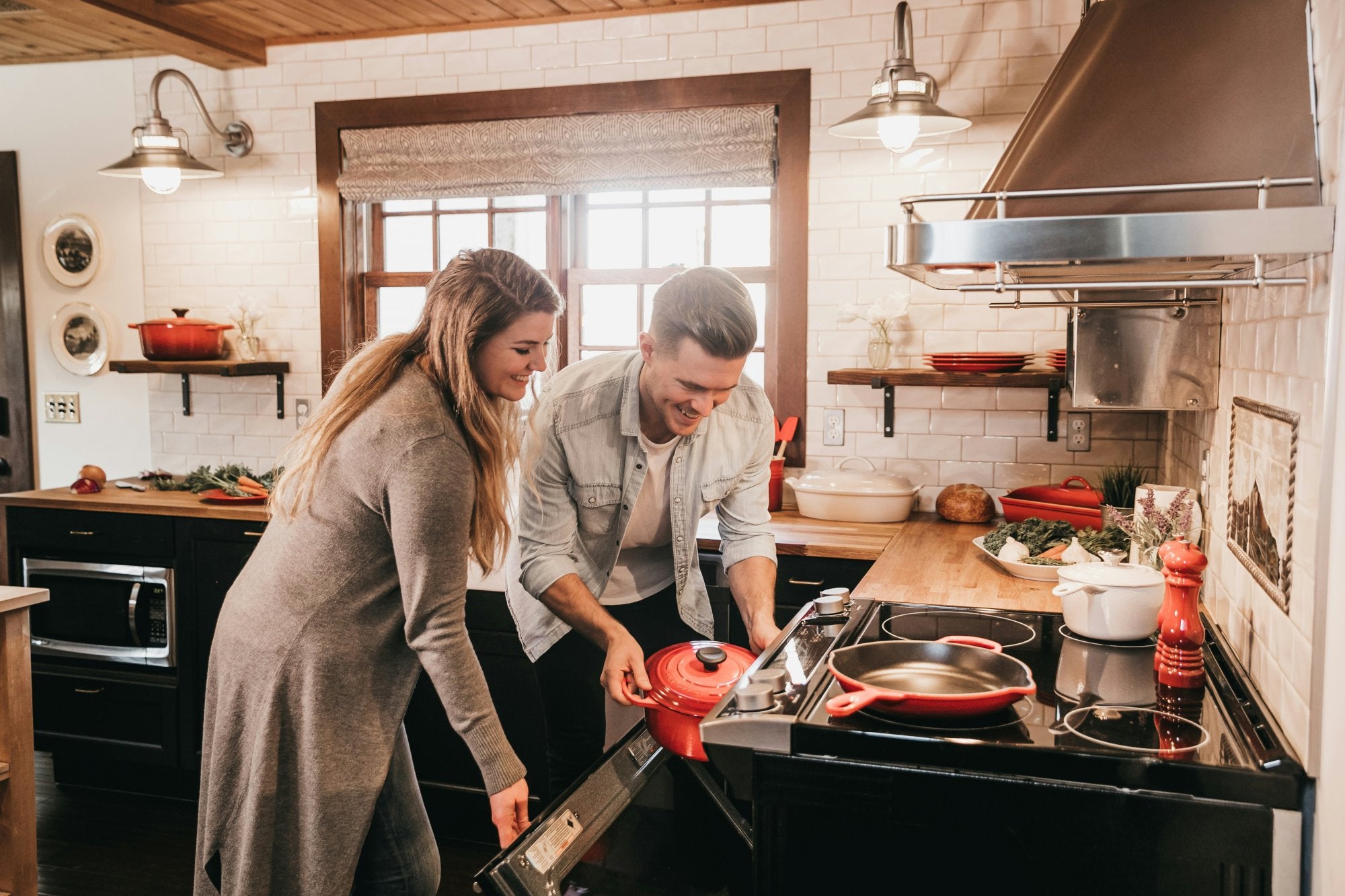 A white male and female place a pot into their electric oven in a cosily lit kitchen scene