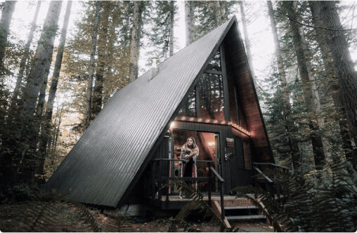 A young, white female stands on the veranda of an illuminated A-frame cabin in a woodland scene